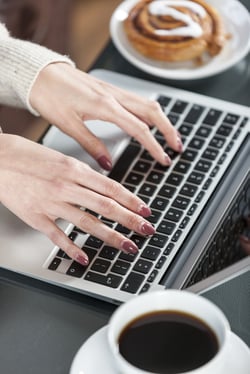 Woman's hands typing on a laptop keyboard with coffee and fresh cake